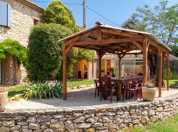 A wooden pergola with a table and chairs, surrounded by a stone wall and greenery.
