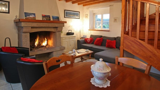 A cozy living room with a fireplace, wooden beams, a sofa with red cushions, and a dining table with a decorative pot.
