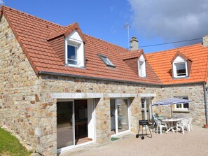 A stone building with a red-tiled roof, a patio with a grill and chairs, and a clear sky.