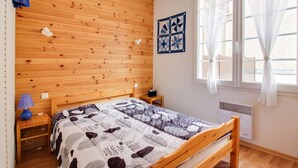 Une chambre avec une tête de lit en bois, un lit recouvert d’une literie à motifs, une table de chevet, une lampe fixée au mur, une fenêtre ornée de rideaux légers et une œuvre d’art matelassée accrochée au mur.