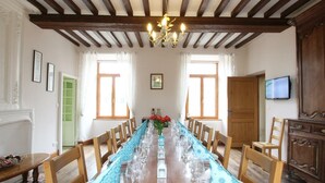 A dining room with a long wooden table set for a meal, flanked by wooden chairs, a chandelier, and a television on the wall. - Charming family cottage, 12 beds, garden and terrace (La Ronde-Haye)