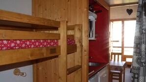 Un lit superposé en bois avec une couvre-lit à motifs rouges et blancs, un coin cuisine avec un mur rouge et des placards blancs, et un coin repas avec une table et des chaises.