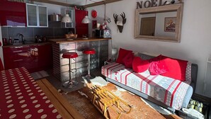 A cozy living room with a sofa, a wooden table, and a kitchen area with a red cabinet and a hanging light fixture. - D104 - 42 m2 - 2P+cab - 4/6 pers (Prapoutel)