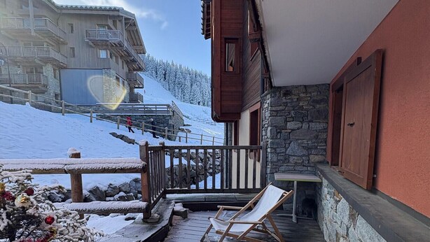 A wooden deck with two lounge chairs, a stone wall, and a view of a snowy mountain and a building in the background. - D104 - 42 m2 - 2P+cab - 4/6 pers (Prapoutel)