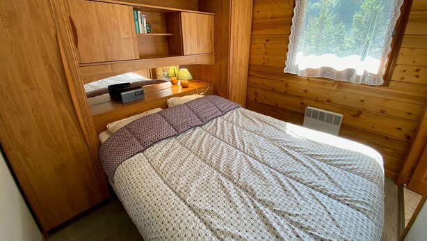 Acogedora habitación tipo cabaña con cama, estanterías de madera, una ventana con cortinas de encaje y una mesita con lámpara.