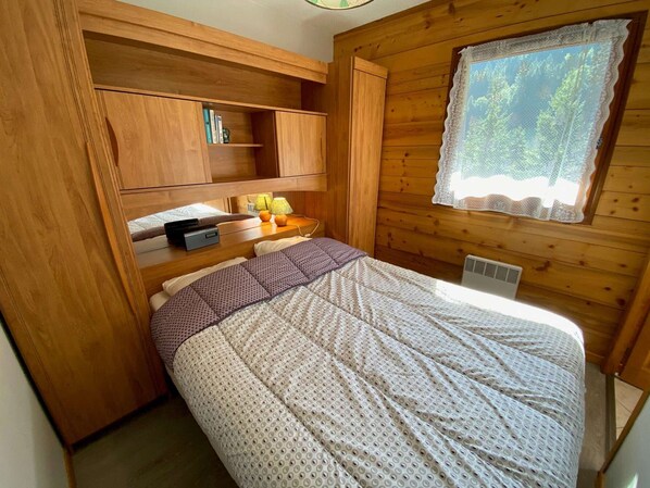 A cozy cabin room with a bed, wooden shelves, a window with lace curtains, and a small table with a lamp.