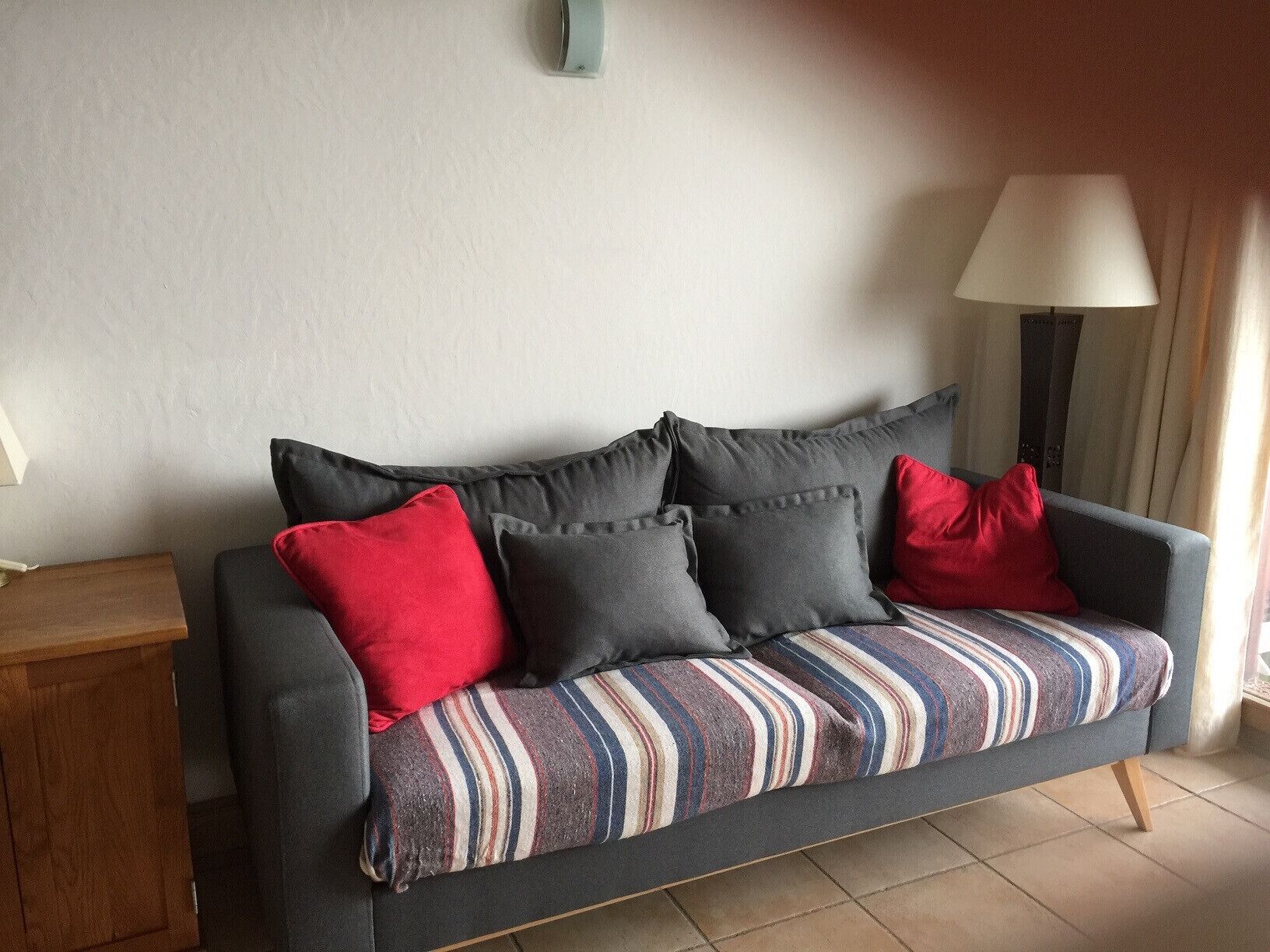 Un sofá gris con almohadones a rayas y cojines decorativos rojos, ubicado frente a una pared blanca con una lámpara y cortinas.
