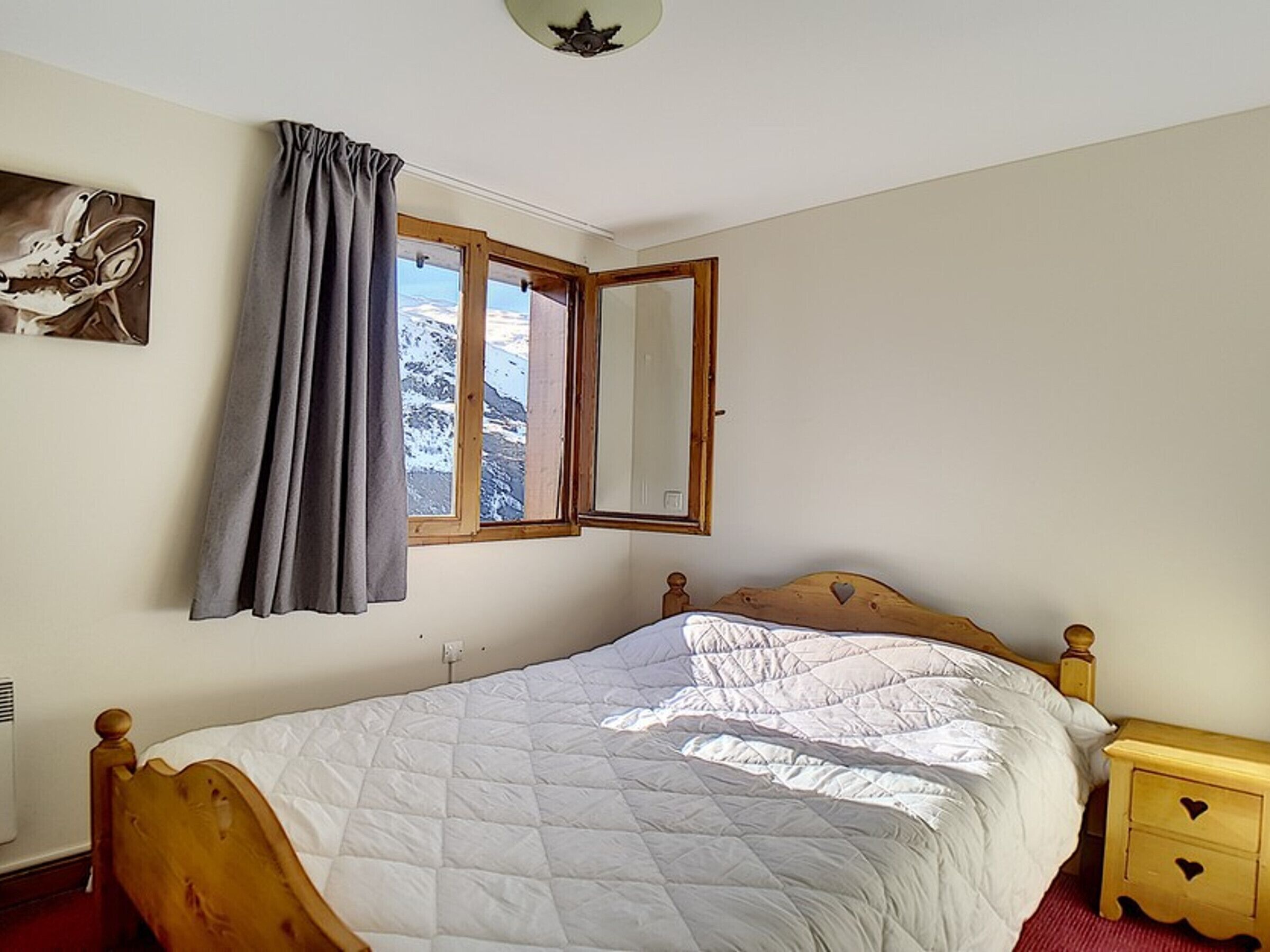 A bedroom with a bed, a window with curtains, a bedside table, and a wall-mounted mirror.