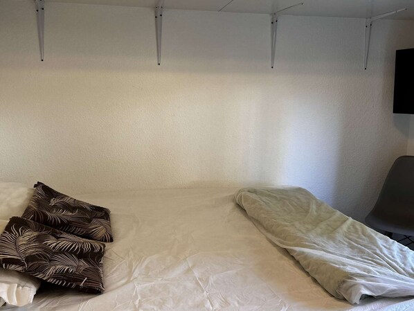 A single bed with a white mattress and a brown patterned pillow, a grey chair, and a white wall with a mounted television. - Charming T2 Cabin with terrace and pool in Port Barcarès (Port Barcarès)