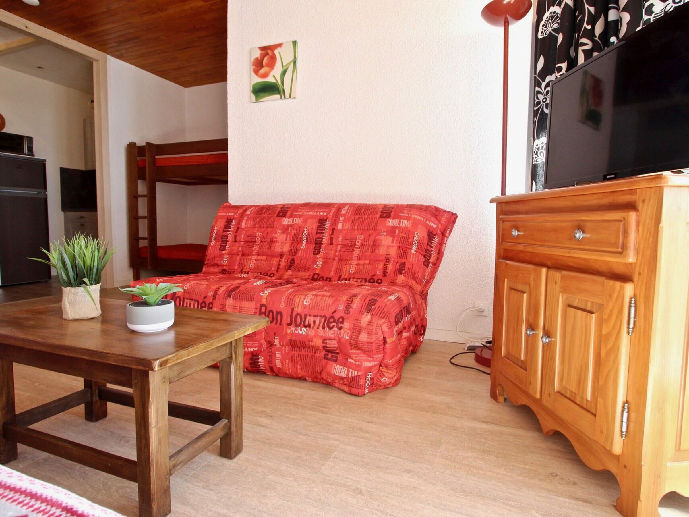 A cozy living room with a wooden table, a red patterned sofa, a wooden cabinet, and a television.