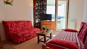 Ein Zimmer mit einem roten, gemusterten Sofa, einem Holztisch mit einer Pflanze und einem Bett mit roter Decke.