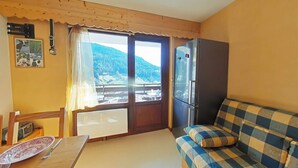 Une pièce avec un plafond en bois, un canapé à carreaux, un réfrigérateur, une table avec une assiette et des couverts, une fenêtre donnant sur les montagnes et un tableau accroché au mur.