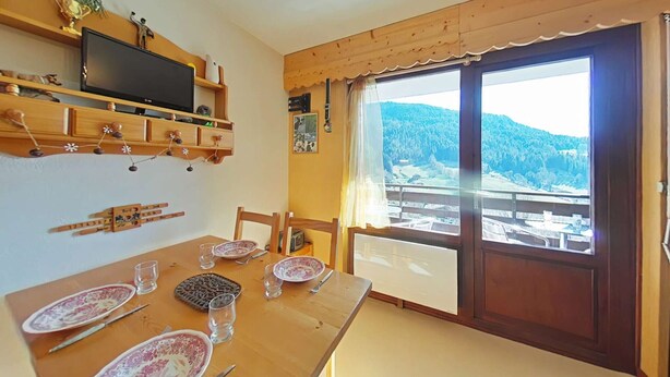 Un coin repas avec une table en bois dressée pour quatre personnes, un téléviseur posé sur une étagère et une grande fenêtre offrant une vue panoramique.