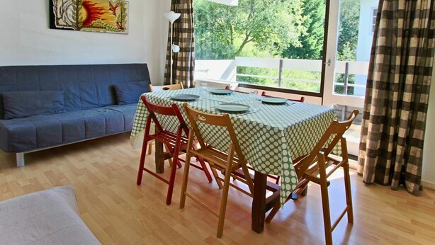 A dining area with a table set for four, a blue sofa, and a large window with a view of greenery. - Cozy studio for 3 near slopes and shops (Chamrousse)