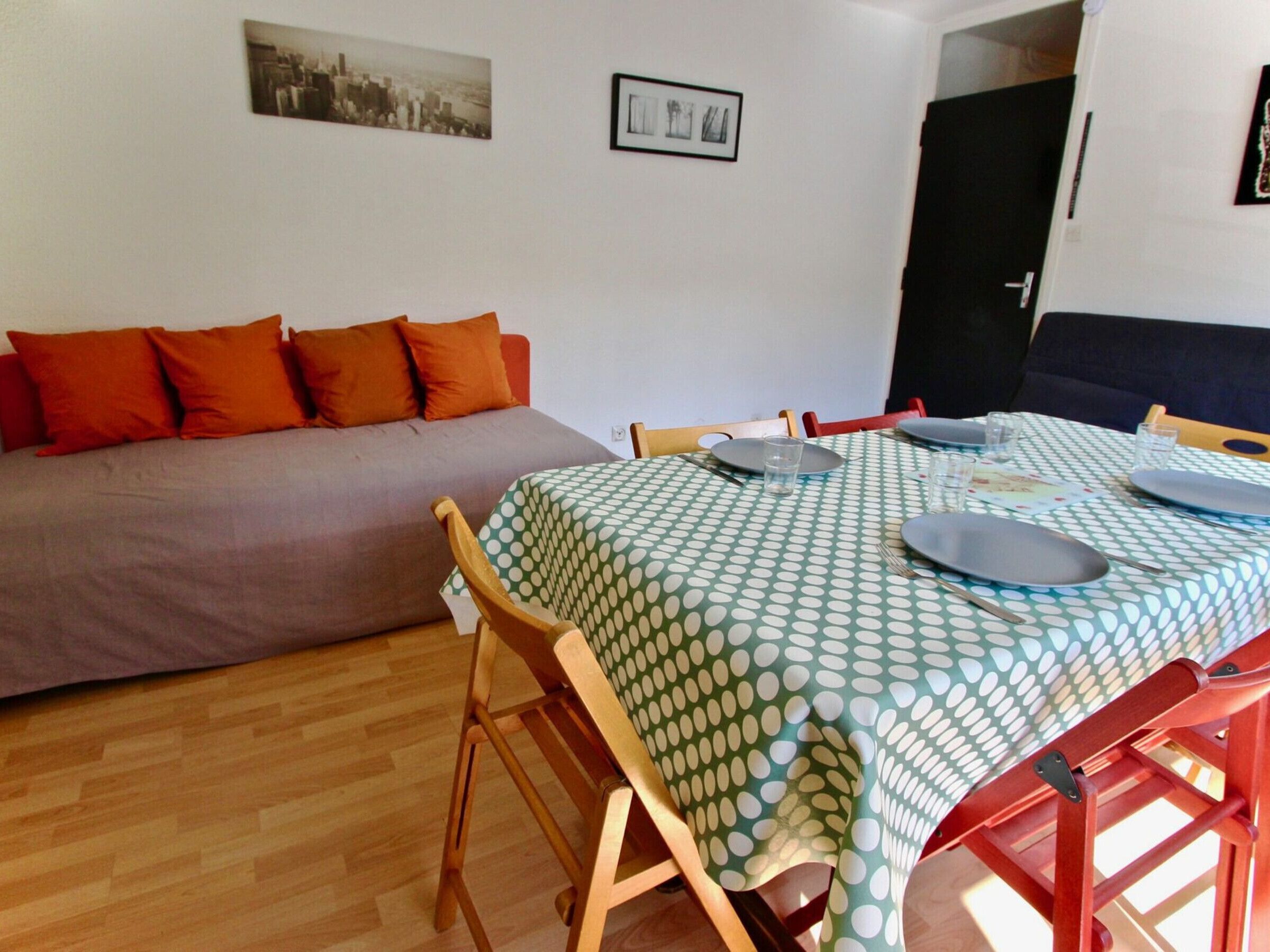 A dining area with a table set for four, a sofa with orange cushions, and a cityscape picture on the wall.