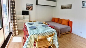 A compact living space with a dining table set for four, a sofa with orange cushions, and a television on a white shelf. - Cozy studio for 3 near slopes and shops (Chamrousse)