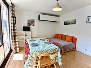 A compact living space with a dining table set for four, a sofa with orange cushions, and a television on a white shelf.