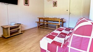 Una habitación con piso de madera, una mesa de comedor con bancos, un sofá, un televisor montado en la pared y dos cuadros enmarcados.