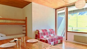 Una habitación acogedora con una cama litera de madera, una mesa de comedor con platos y vasos, una manta escocesa en una silla y una ventana con vistas a la vegetación.