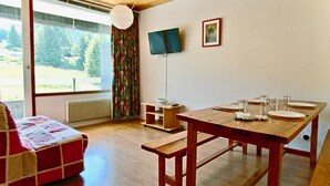 Una habitación acogedora con una mesa de comedor de madera dispuesta para dos, un sofá rojo y blanco, un televisor en la pared y una ventana con vistas a árboles.