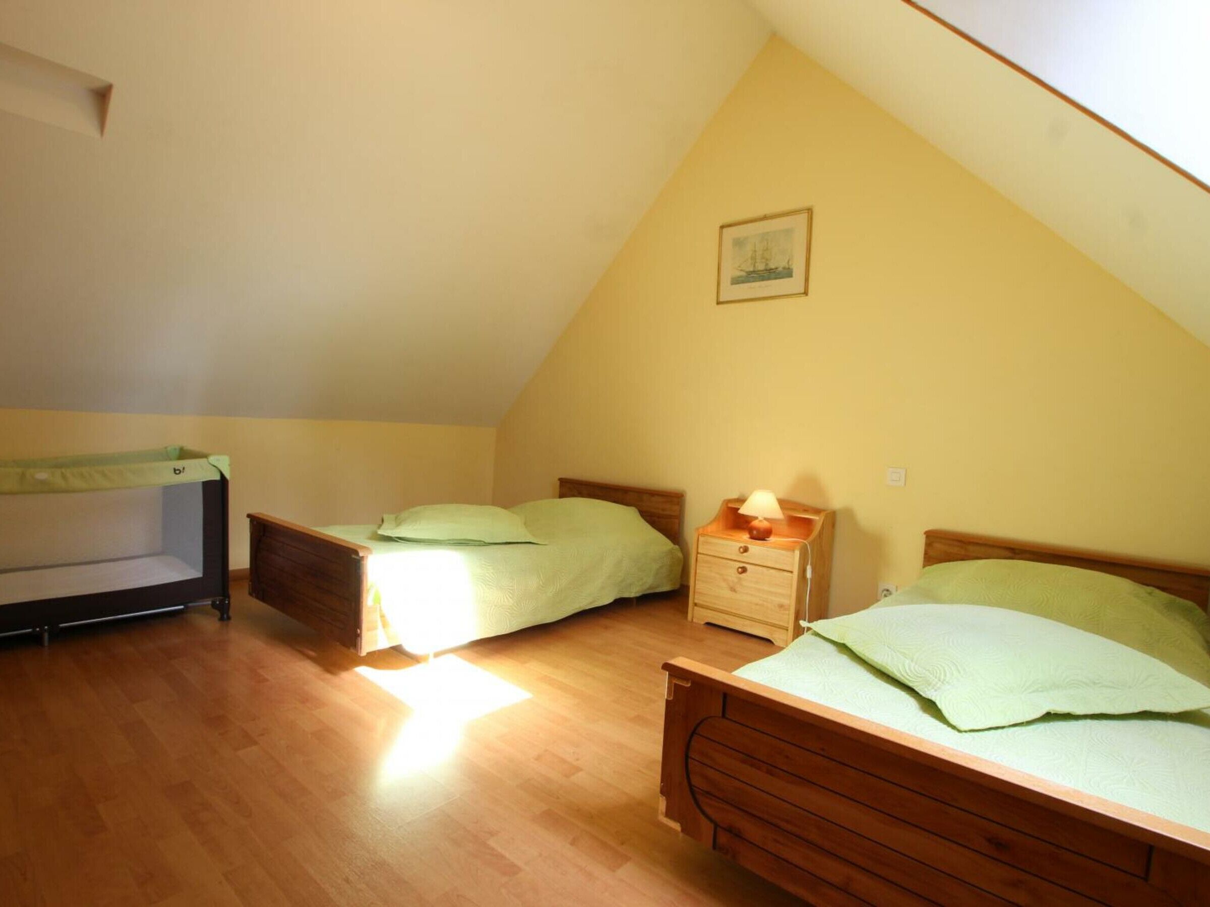 Une petite chambre mansardée triangulaire, avec deux lits en bois, une table de chevet et un berceau.