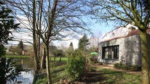 A modern house with a large window, surrounded by a garden and a body of water.