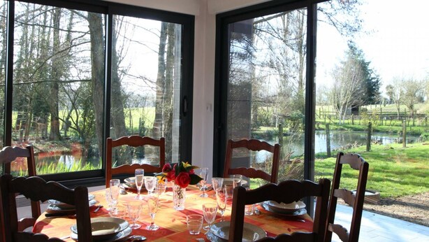 A dining table set for six with chairs, large windows offering a view of a pond and trees, and a vase with flowers on the table.