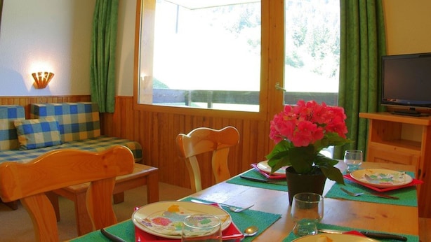 Una habitación acogedora con un sofá de madera, una mesa de comedor con un florero y un televisor sobre un soporte.