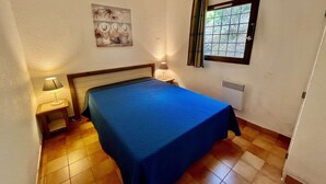 A bedroom with a bed, a nightstand, a lamp, a window with curtains, and a wall-mounted radiator. - Apartment with Pool and Terrace 300m from the Beach (La Croix-Valmer)