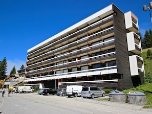 A multi-story building with balconies, parking area, and a small structure with a sign.
