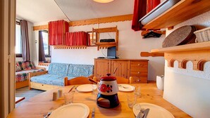 Et hyggeligt rum med et træbord dækket til fire personer, en sofa med mønstrede puder, et træskab og et fjernsyn på et træstel.
