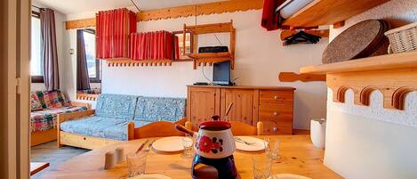 A cozy room with a wooden dining table set for four, a sofa with patterned cushions, a wooden cabinet, and a television on a wooden stand.