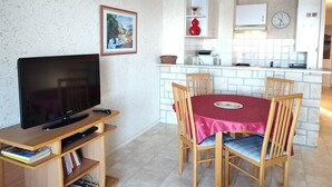 A compact living space with a TV on a wooden stand, a dining table with four chairs, a kitchen area with a clock, and a framed picture on the wall.
