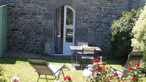 A stone building with a small patio area featuring a table and two chairs, surrounded by a garden with flowers. - Charming studio at the Manor with private garden and WiFi (Baubigny)