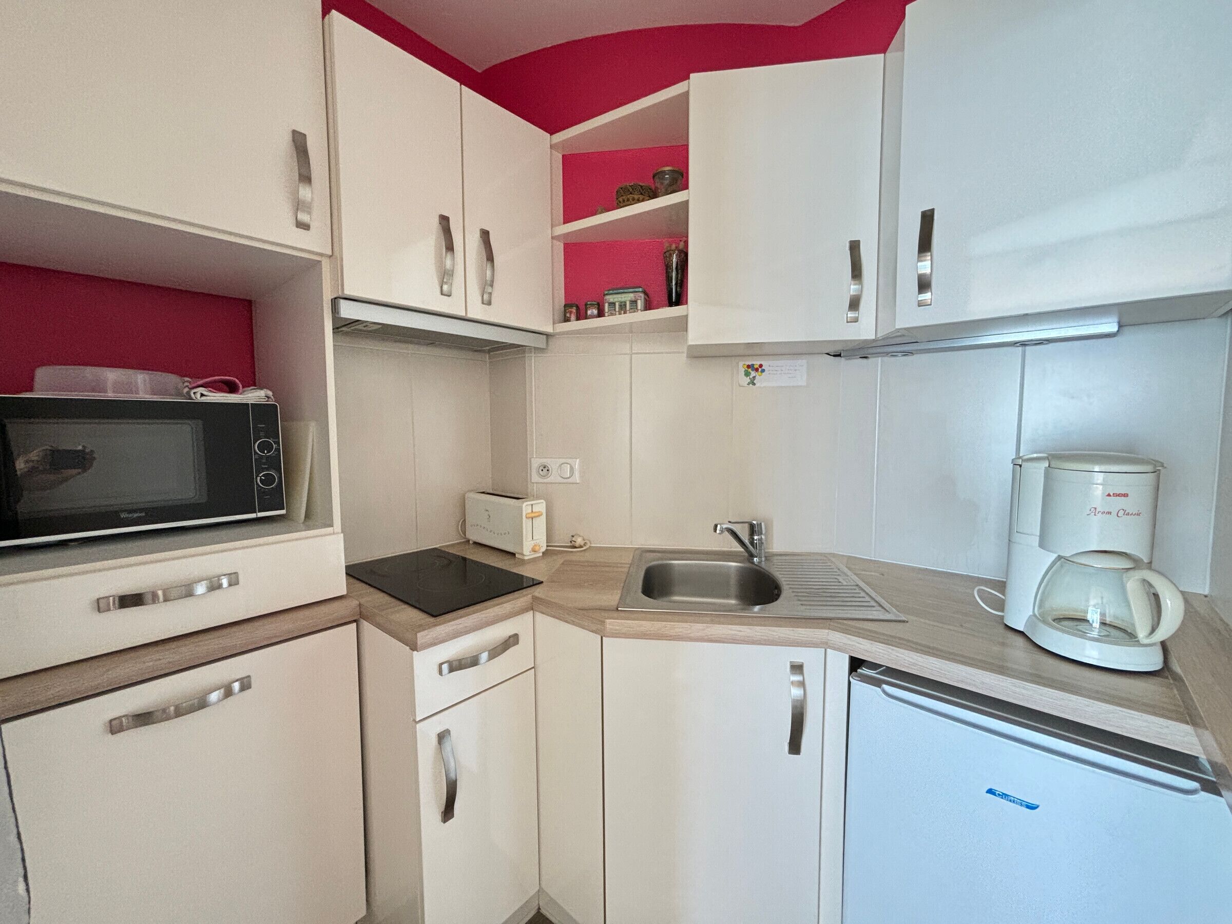 Een kleine keuken met witte kastjes, een magnetron, een broodrooster, een spoelbak en een koffiezetapparaat.