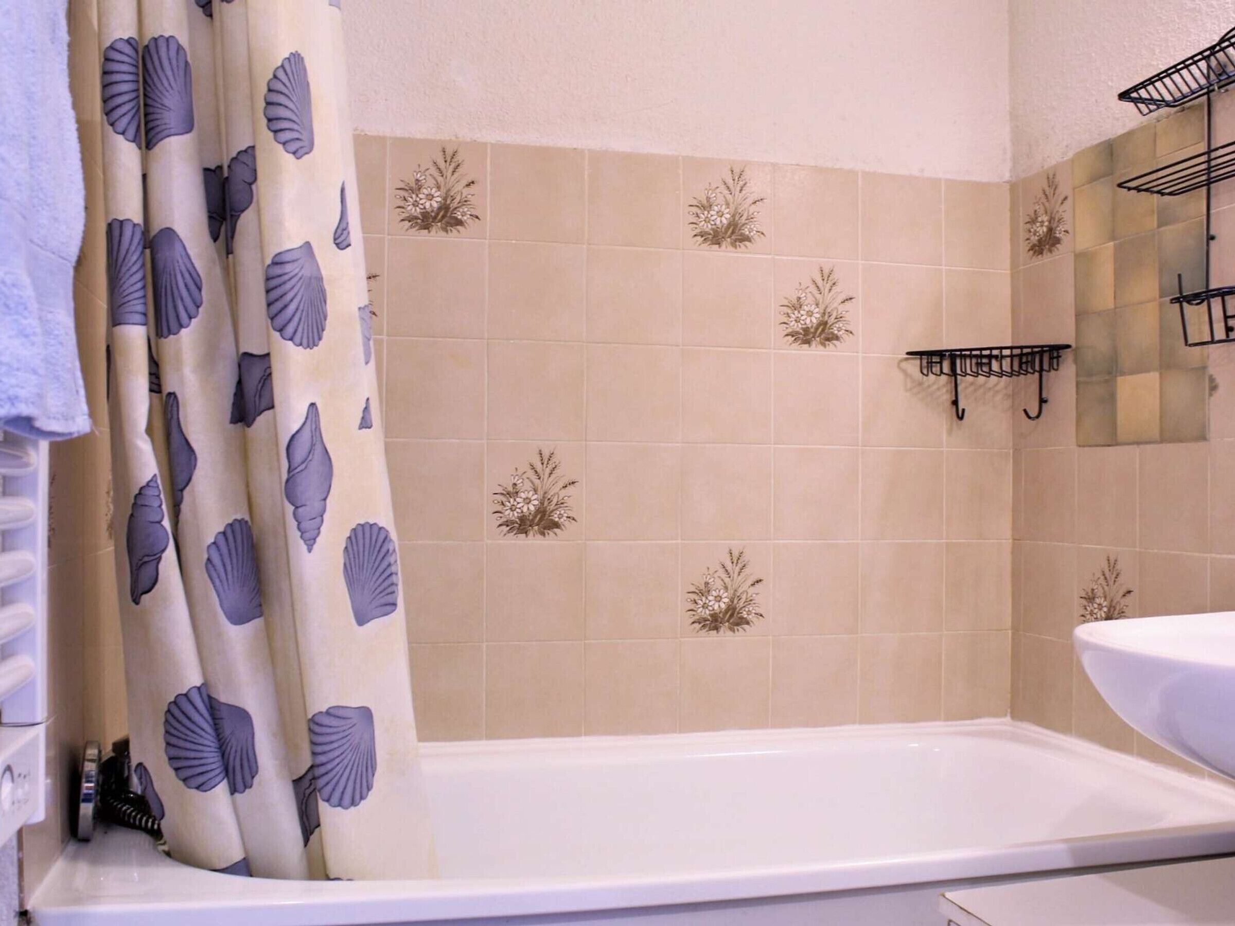 A bathroom with a bathtub, a shower curtain with a shell pattern, and beige tiles with floral designs.
