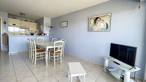 A modern living room with a dining area, a kitchen in the background, and a large TV on a stand. - 1 Bedroom Apartment + Cabin with Sea & Dune View, 3rd Floor with Elevator (Cucq-Stella Plage)