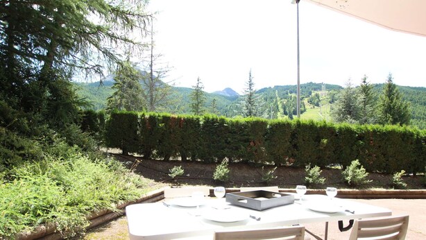 An outdoor dining area with a table and chairs, surrounded by greenery and a view of mountains.