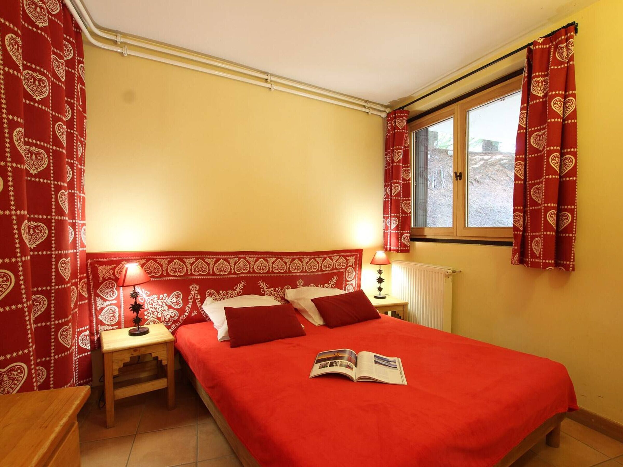 Une chambre à coucher avec une couvre-lit rouge, deux lampes de chevet, une table de chevet en bois et une fenêtre ornée de rideaux rouges.