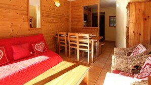 A cozy wooden cabin interior with a red sofa, a dining table with chairs, and a wicker chair.