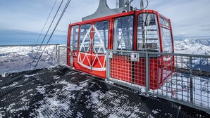 Un teleférico rojo con un logo triangular, suspendido por cables, con vistas a un paisaje montañoso nevado.