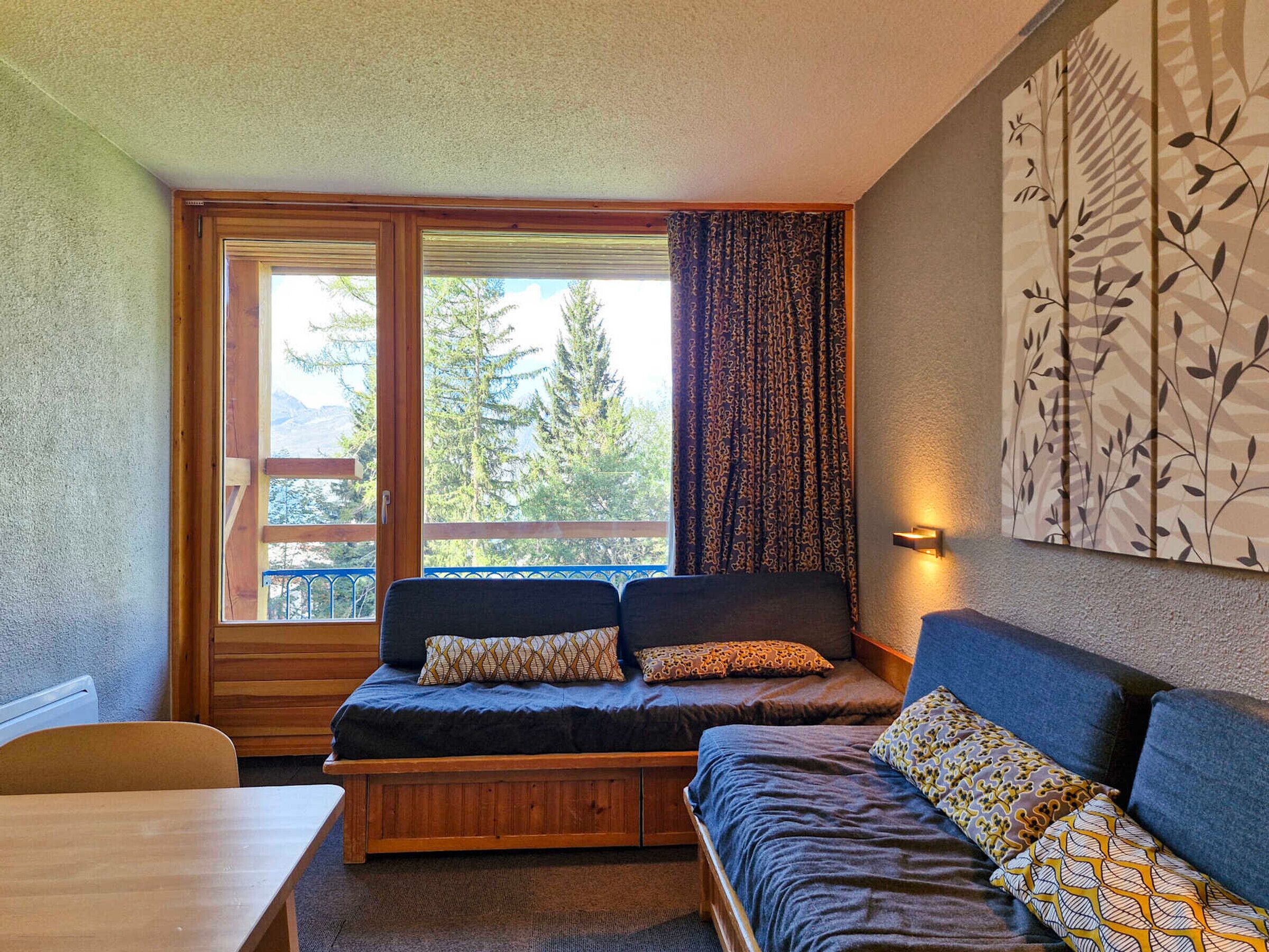 A cozy room with a sofa, a wooden bench, and a table. There is a large window with curtains, and a wall-mounted light fixture.