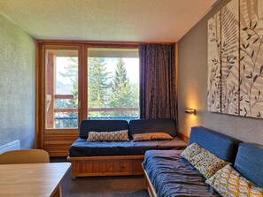 A cozy room with a sofa, a wooden bench, and a table. There is a large window with curtains, and a wall-mounted light fixture.