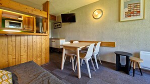 A compact room with a wooden kitchenette, a dining table with chairs, a small stool, and a television mounted on the wall. - Ski-in/out studio for 5, Les Arcs 1800, pets allowed (Les Arcs 1800)