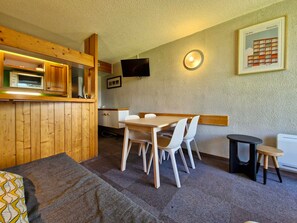 A compact room with a wooden kitchenette, a dining table with chairs, a small stool, and a television mounted on the wall. - Ski-in/out studio for 5, Les Arcs 1800, pets allowed (Les Arcs 1800)