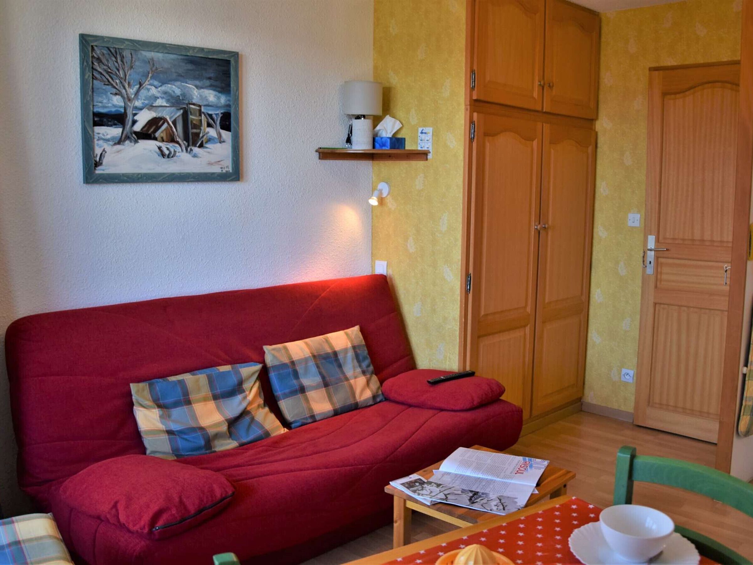 Een gezellige kamer met een rode bank, geruite kussens, een klein tafeltje met een schaal en een boek, en een houten garderobe.