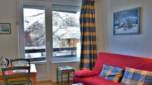 A cozy living room with a red sofa, a small wooden table, a green chair, and a window with a view of a snowy landscape. - Mountain Studio, 4 Sleeps, Ski-In/Ski-Out, Free Parking, Risoul 1850 (Risoul)