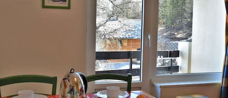 Uma área de refeições com uma mesa posta para dois, uma chaleira, uma caixa de cereal e uma janela com vista para árvores e um edifício.