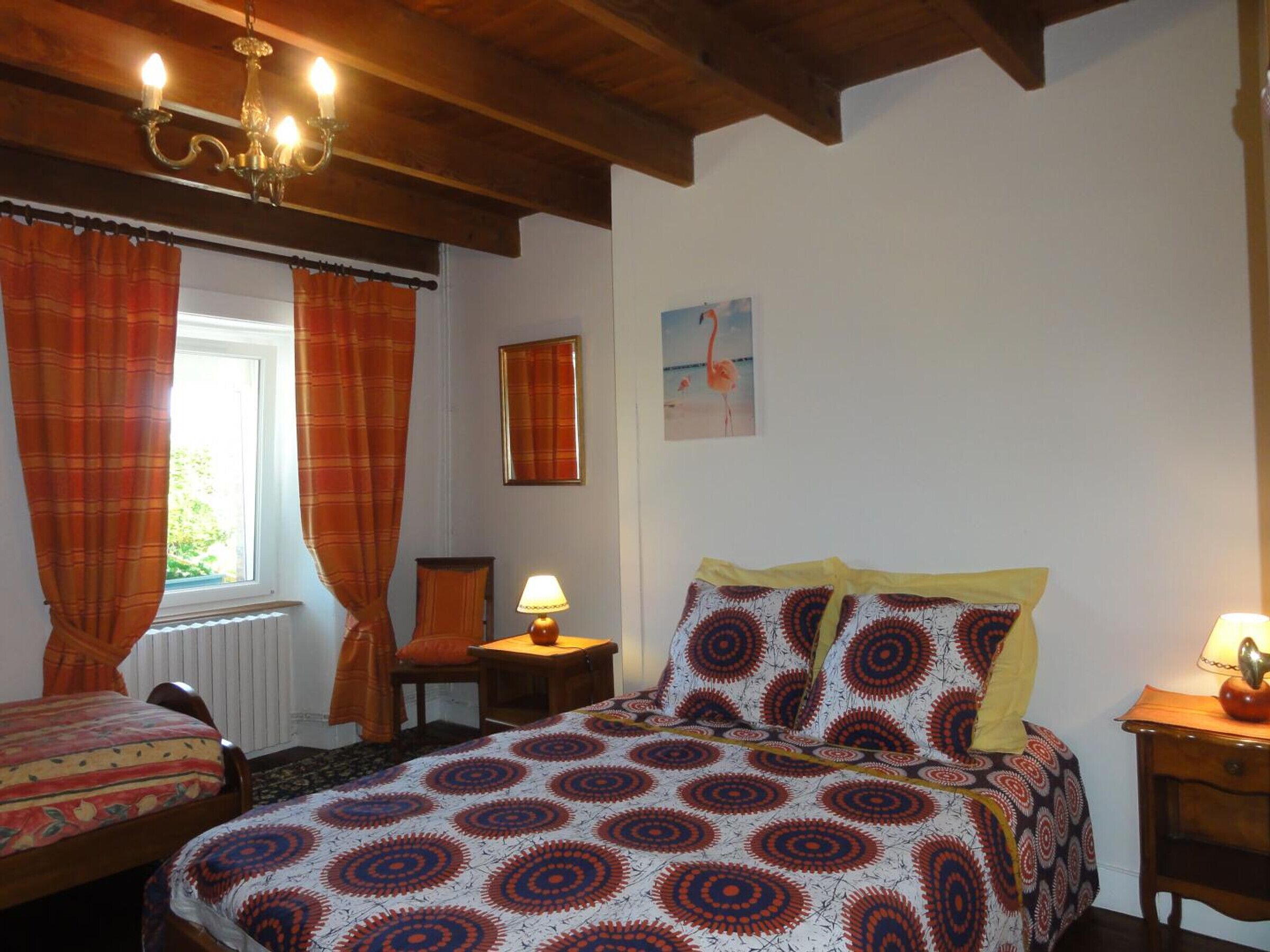 A bedroom with a bed, two bedside tables, a chair, a window with curtains, a wall-mounted picture, and a chandelier.