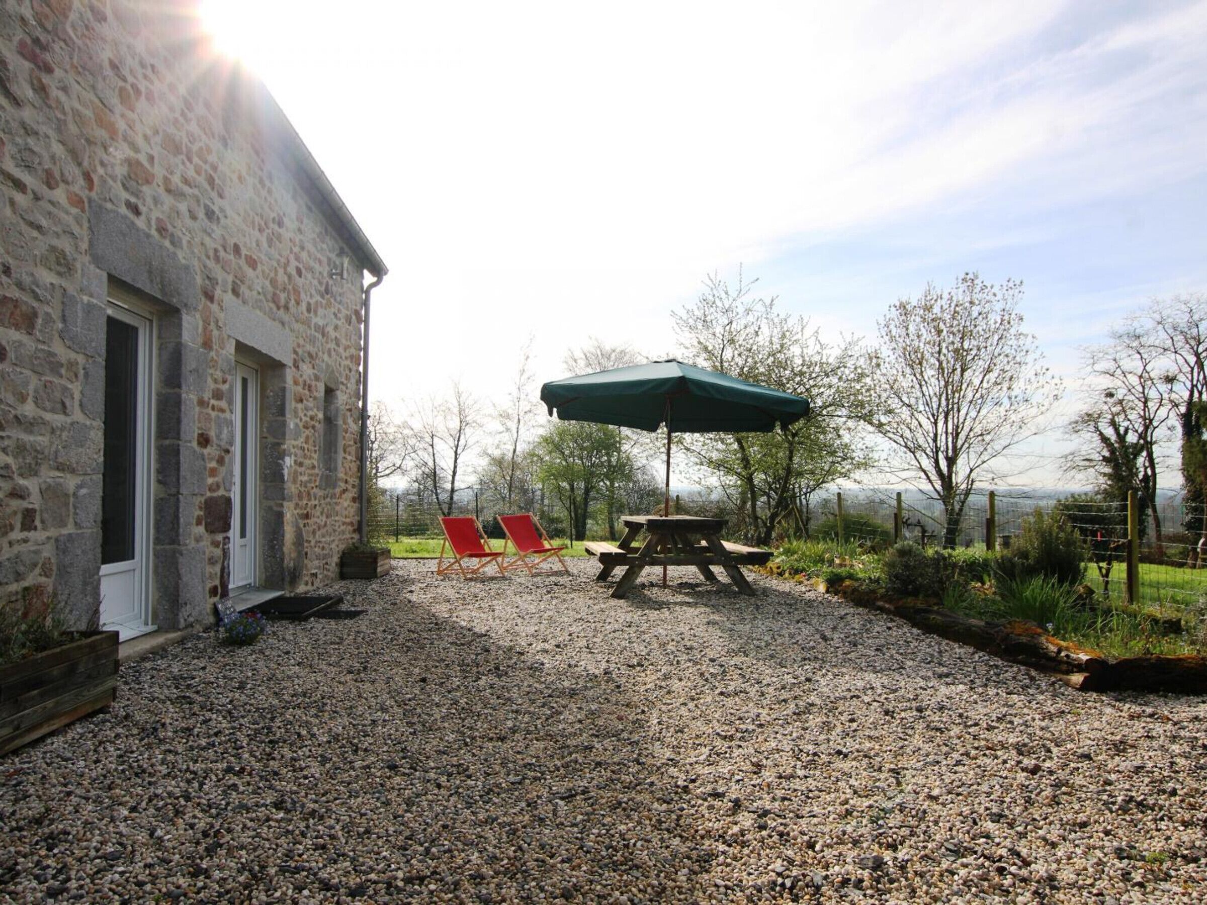 A stone building with a gravel patio, a picnic table, and red chairs under a green umbrella.